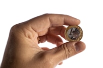 A hand is holding a Brazilian one real coin between the thumb and index finger. The coin features a metallic design with a mix of silver and gold colors, and the words '1 REAL' are visible.