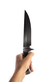 A hand is gripping a large knife with intricate, ornamental metalwork on the handle against a plain white background. The knife has a long, sharp blade.