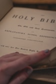 An open page of the Holy Bible, featuring titles and text in an old-style font. A hand with painted fingernails gently turns the page.