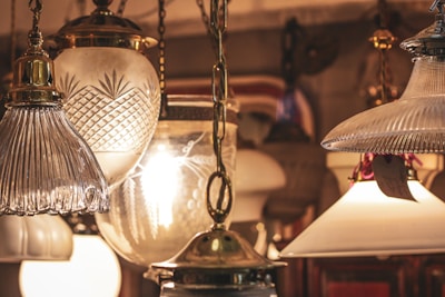 A collection of vintage-style hanging lamps with various glass designs, including ribbed, cut, and frosted glass. Warm light emanates from the bulbs inside, creating an antique and cozy ambiance. The lamps are suspended from ornate brass chains.