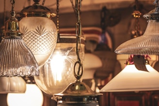 A collection of vintage-style hanging lamps with various glass designs, including ribbed, cut, and frosted glass. Warm light emanates from the bulbs inside, creating an antique and cozy ambiance. The lamps are suspended from ornate brass chains.