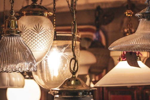 A collection of vintage-style hanging lamps with various glass designs, including ribbed, cut, and frosted glass. Warm light emanates from the bulbs inside, creating an antique and cozy ambiance. The lamps are suspended from ornate brass chains.