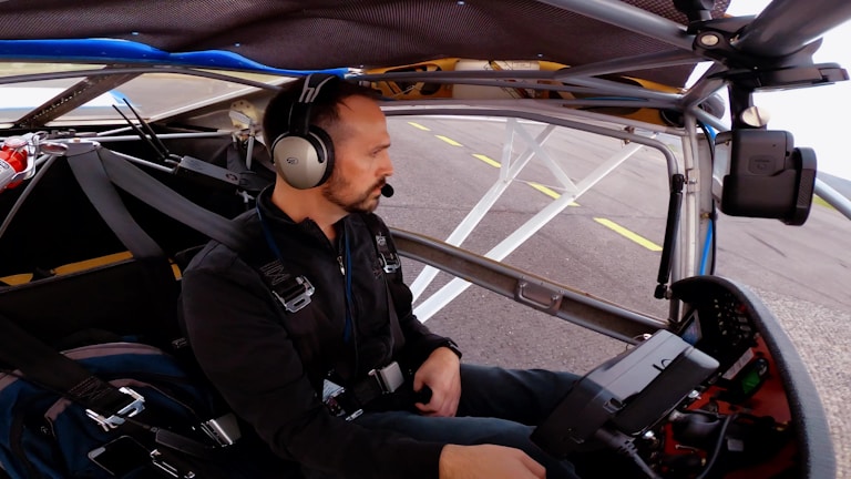 A person wearing a headset is seated inside a small aircraft cockpit. They are surrounded by various controls and equipment, with the aircraft's structure visible around them. The view outside shows part of a runway and the sky.