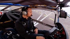 A person wearing a headset is seated inside a small aircraft cockpit. They are surrounded by various controls and equipment, with the aircraft's structure visible around them. The view outside shows part of a runway and the sky.