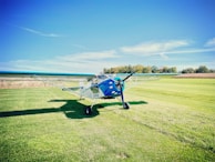 A small, single-engine airplane sits on a grassy field under a clear blue sky. The plane has a blue and white color scheme, with the name 'Kitfox' visible on its body. In the background, there is a stretch of trees and shrubs separating the field from the horizon.