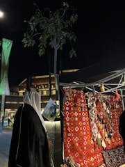 A night scene featuring a traditional market stall with colorful woven textiles and tassels on display. A person wearing traditional Middle Eastern attire stands nearby, and lush green foliage can be seen above. Modern architecture, lit with green lights, forms the backdrop.