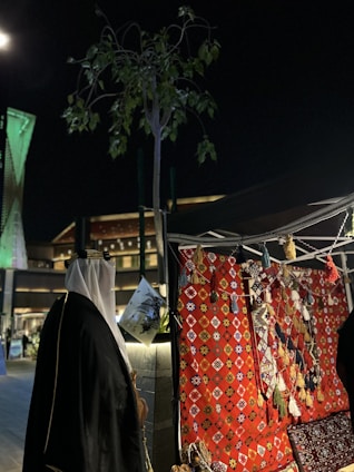 A night scene featuring a traditional market stall with colorful woven textiles and tassels on display. A person wearing traditional Middle Eastern attire stands nearby, and lush green foliage can be seen above. Modern architecture, lit with green lights, forms the backdrop.