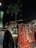 A night scene featuring a traditional market stall with colorful woven textiles and tassels on display. A person wearing traditional Middle Eastern attire stands nearby, and lush green foliage can be seen above. Modern architecture, lit with green lights, forms the backdrop.
