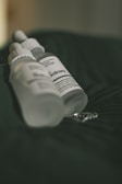 A close-up view of two frosted dropper bottles lying on a dark green fabric. The bottles are labeled with text, including 'The Ordinary,' and appear to be skincare products. The lighting is soft, casting gentle shadows and creating a slightly moody atmosphere.
