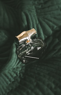 A clear, intricately designed perfume bottle with a golden cap is resting on a dark green, textured fabric. The fabric appears to have a velvety surface with distinct folds and ridges, adding depth to the overall composition.