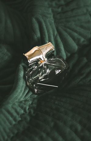 A clear, intricately designed perfume bottle with a golden cap is resting on a dark green, textured fabric. The fabric appears to have a velvety surface with distinct folds and ridges, adding depth to the overall composition.
