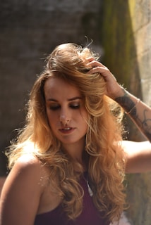 A woman with long, curly blonde hair is positioned in a softly lit environment, touching her hair with one hand. She has a contemplative expression and is wearing a sleeveless top. The background features a rustic, textured wall with hints of greenery.