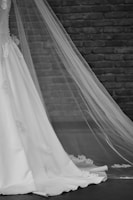 A white wedding dress with intricate lace details and a long, flowing veil. The background consists of a brick wall with a rustic appearance. The focus is on the lower part of the dress and the lengthy veil extending to the ground.