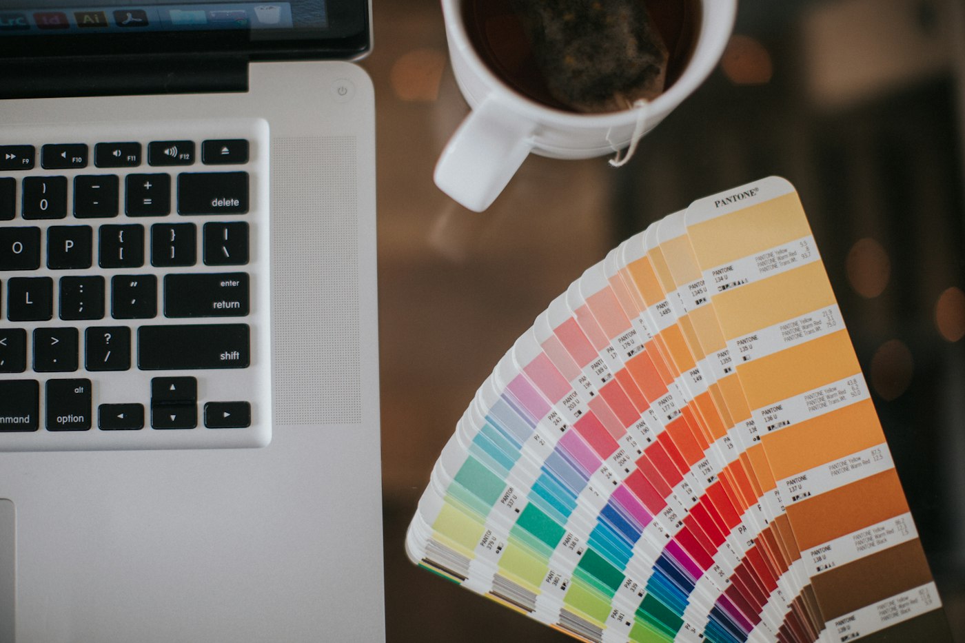 A fan of printed color swatches sits beside a laptop and a cup on a dark glass desk.