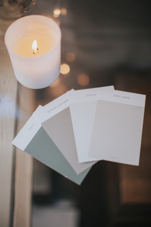 A lit white candle is placed on a reflective surface next to several paint color swatches. The candle creates a warm, cozy ambiance with its soft glow, while the swatches display muted, neutral colors with names like Eden and Shiplap.
