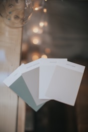 Several paint color swatches are fanned out on a surface, with colors ranging from greenish-grey to off-white. In the background, there is a soft focus of string lights creating a bokeh effect, adding a warm ambiance.