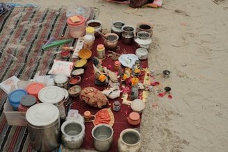 A collection of various containers and ritual items are arranged on a red cloth placed on sand. There are multiple stainless steel and plastic containers, along with items such as combs, incense sticks, small bowls with colored powders, and decorative objects like marigold flowers. The scene suggests a setup for a traditional ritual or offering.