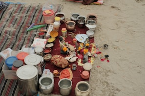 A collection of various containers and ritual items are arranged on a red cloth placed on sand. There are multiple stainless steel and plastic containers, along with items such as combs, incense sticks, small bowls with colored powders, and decorative objects like marigold flowers. The scene suggests a setup for a traditional ritual or offering.