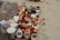 A collection of various containers and ritual items are arranged on a red cloth placed on sand. There are multiple stainless steel and plastic containers, along with items such as combs, incense sticks, small bowls with colored powders, and decorative objects like marigold flowers. The scene suggests a setup for a traditional ritual or offering.