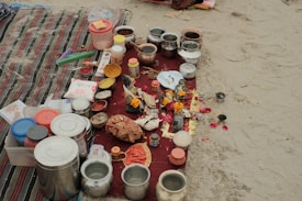 A collection of various containers and ritual items are arranged on a red cloth placed on sand. There are multiple stainless steel and plastic containers, along with items such as combs, incense sticks, small bowls with colored powders, and decorative objects like marigold flowers. The scene suggests a setup for a traditional ritual or offering.