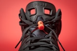 A close-up view of a black sneaker with intricate lace details and a prominent orange lace lock. The background is a solid red color, making the sneaker's design stand out. The shoe features distinctive patterns with visible branding elements on the tongue.