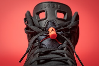 A close-up view of a black sneaker with intricate lace details and a prominent orange lace lock. The background is a solid red color, making the sneaker's design stand out. The shoe features distinctive patterns with visible branding elements on the tongue.