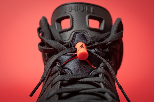 A close-up view of a black sneaker with intricate lace details and a prominent orange lace lock. The background is a solid red color, making the sneaker's design stand out. The shoe features distinctive patterns with visible branding elements on the tongue.