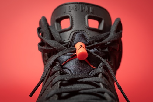 A close-up view of a black sneaker with intricate lace details and a prominent orange lace lock. The background is a solid red color, making the sneaker's design stand out. The shoe features distinctive patterns with visible branding elements on the tongue.