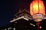 A traditional red lantern with Chinese characters is prominently displayed, glowing warmly against the night. In the background, there is the blurred silhouette of a traditional East Asian architectural structure, suggesting a serene and culturally rich setting.