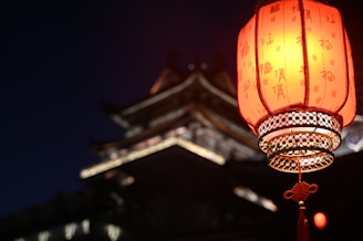 A traditional red lantern with Chinese characters is prominently displayed, glowing warmly against the night. In the background, there is the blurred silhouette of a traditional East Asian architectural structure, suggesting a serene and culturally rich setting.