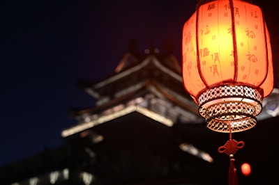 A traditional red lantern with Chinese characters is prominently displayed, glowing warmly against the night. In the background, there is the blurred silhouette of a traditional East Asian architectural structure, suggesting a serene and culturally rich setting.