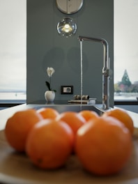 A modern kitchen setting with a focus on a sleek chrome faucet running a thin stream of water. In the foreground, a wooden bowl holds several oranges, while a single white vase with a flower decorates the countertop. A pendant light hangs above, casting a subtle glow. Views of a lake and trees are visible through large windows, providing a serene backdrop.