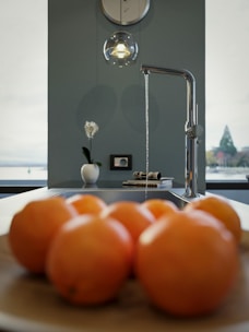 A modern kitchen setting with a focus on a sleek chrome faucet running a thin stream of water. In the foreground, a wooden bowl holds several oranges, while a single white vase with a flower decorates the countertop. A pendant light hangs above, casting a subtle glow. Views of a lake and trees are visible through large windows, providing a serene backdrop.