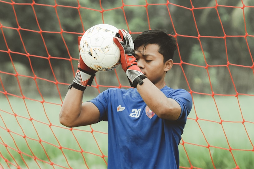 A person wearing a blue sports jersey and red gloves holds a soccer ball in front of their face. They stand in front of a red soccer net, with a blurred green field in the background. The individual appears focused and concentrated.