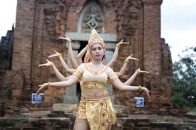A dancer wearing traditional attire poses in front of an ancient brick structure. The costume is ornate, with gold and red details, and the dancer is positioned to create an illusion of having multiple arms, reminiscent of cultural or mythological representations. The background features a historical temple with intricate carvings.