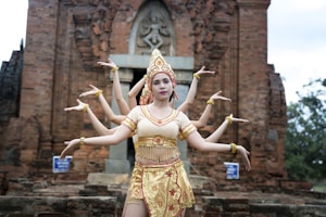A dancer wearing traditional attire poses in front of an ancient brick structure. The costume is ornate, with gold and red details, and the dancer is positioned to create an illusion of having multiple arms, reminiscent of cultural or mythological representations. The background features a historical temple with intricate carvings.