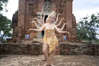 A group of dancers wearing traditional attire is performing in front of an ancient brick temple. The setting is outdoors with trees in the background, and the dancers are elegantly posing with their arms extended in unison.