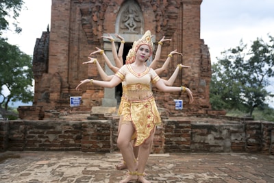 A group of dancers wearing traditional attire is performing in front of an ancient brick temple. The setting is outdoors with trees in the background, and the dancers are elegantly posing with their arms extended in unison.
