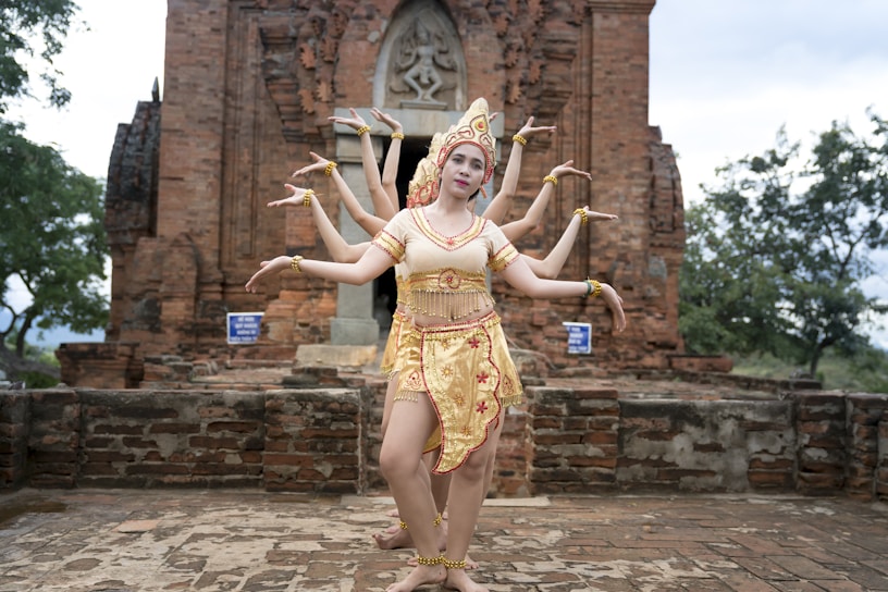 A group of dancers wearing traditional attire is performing in front of an ancient brick temple. The setting is outdoors with trees in the background, and the dancers are elegantly posing with their arms extended in unison.