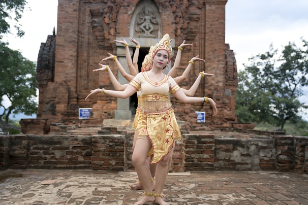 A group of dancers wearing traditional attire is performing in front of an ancient brick temple. The setting is outdoors with trees in the background, and the dancers are elegantly posing with their arms extended in unison.