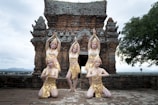 Five dancers perform in front of an ancient stone temple. They wear traditional golden costumes with ornate headpieces and pose elegantly with arms raised. The temple architecture features intricate carvings and a large central archway. The setting is outdoors, with a view of the landscape in the background, including some trees and distant mountains.