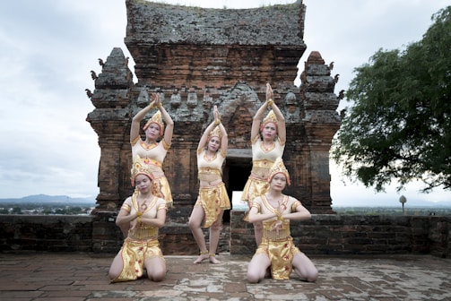 Five dancers perform in front of an ancient stone temple. They wear traditional golden costumes with ornate headpieces and pose elegantly with arms raised. The temple architecture features intricate carvings and a large central archway. The setting is outdoors, with a view of the landscape in the background, including some trees and distant mountains.