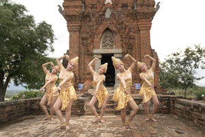 Five dancers in traditional attire pose in front of an ancient, intricately designed brick temple. The dancers' costumes are elaborate, consisting of gold and yellow garments with detailed patterns and headpieces. The temple showcases carvings and an arched entrance, set on a stone platform surrounded by lush greenery.
