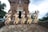 Five people dressed in traditional cultural attire perform a synchronized dance in front of an ancient brick temple with ornate carvings. They wear elaborate headdresses and vibrant costumes with intricate patterns. The setting is outdoors, surrounded by lush greenery and an overcast sky.