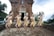 Five people dressed in traditional cultural attire perform a synchronized dance in front of an ancient brick temple with ornate carvings. They wear elaborate headdresses and vibrant costumes with intricate patterns. The setting is outdoors, surrounded by lush greenery and an overcast sky.
