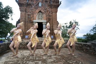 Five people dressed in traditional cultural attire perform a synchronized dance in front of an ancient brick temple with ornate carvings. They wear elaborate headdresses and vibrant costumes with intricate patterns. The setting is outdoors, surrounded by lush greenery and an overcast sky.