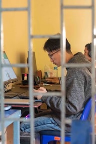 A person wearing glasses is focused on a task at a desk with a computer monitor and various office supplies around. The person is seated on a blue chair, and the view is partially obstructed by metal bars, suggesting a cage-like setting. The environment appears casual and conducive to work.