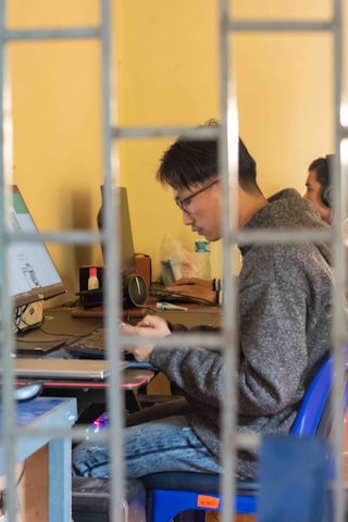 A person wearing glasses is focused on a task at a desk with a computer monitor and various office supplies around. The person is seated on a blue chair, and the view is partially obstructed by metal bars, suggesting a cage-like setting. The environment appears casual and conducive to work.