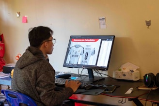 A person wearing a hoodie is seated at a desk, working on a desktop computer. The monitor displays an image editing software showing a sports jersey with the name 'MY FRIEND' and the number 9. The room has a simple decor with a few items on the wall and a packet of tissues on the desk.