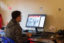A person wearing a hoodie is seated at a desk, working on a desktop computer. The monitor displays an image editing software showing a sports jersey with the name 'MY FRIEND' and the number 9. The room has a simple decor with a few items on the wall and a packet of tissues on the desk.
